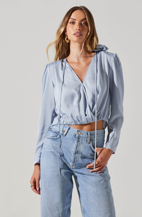 A woman is wearing a light blue satin cropped hoodie with long sleeves and a drawstring hood, paired with high-waisted, light-wash jeans.