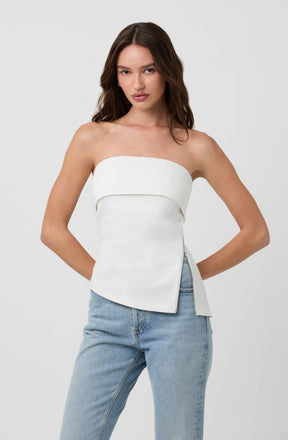 A woman is wearing a strapless white top with a horizontal band across the chest and a fitted, slightly asymmetrical hem. She pairs it with light blue, high-waisted jeans.