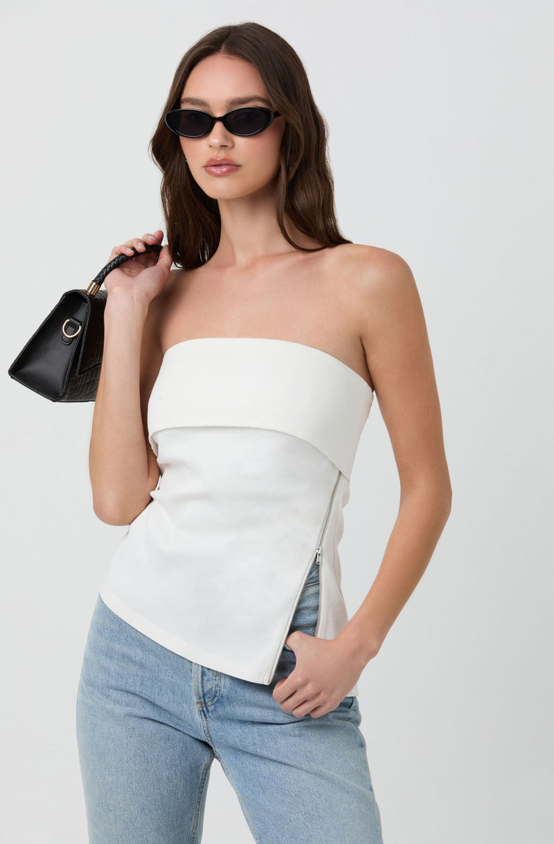 A woman is wearing a strapless white top with a diagonal zipper detail on the side, paired with light blue jeans. She has on black sunglasses and is holding a small black handbag over her shoulder.