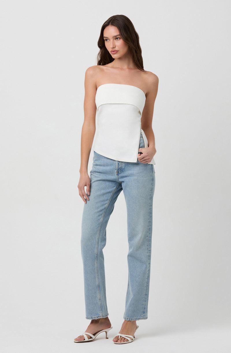 A woman is wearing a strapless white top with a layered design and light blue high-waisted jeans. She has on white heeled sandals with thin straps.