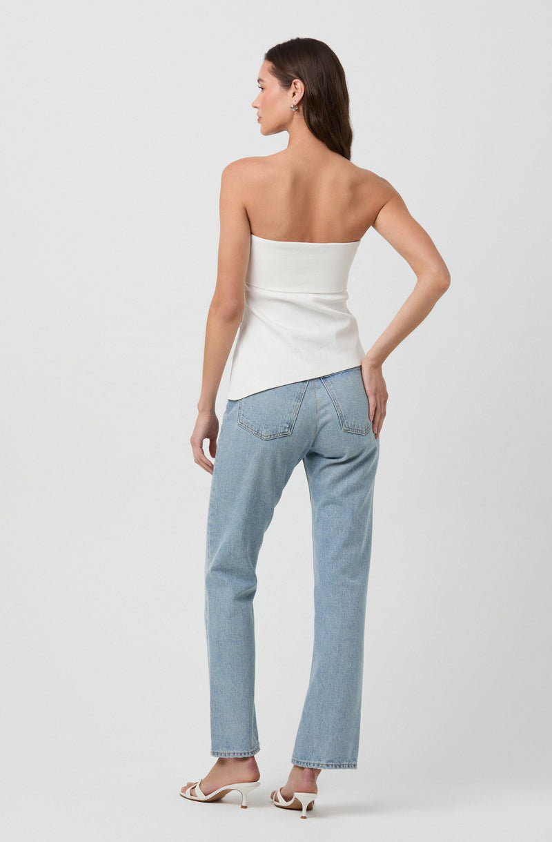 A woman is wearing a strapless white top that is fitted at the waist with a slight asymmetrical hem. She is also wearing light blue high-waisted jeans with a relaxed fit, featuring classic denim stitching and back pockets. She has on white open-toe high heel shoes and is accessorized with small earrings. Her hair is dark and loose, and she is posing with her back turned to the camera, her right hand resting on her hip.