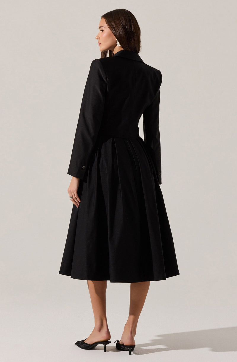 A woman is wearing a black blazer with long sleeves and a fitted waist, paired with a matching black midi-length skirt that has pleats and a full silhouette. She is also wearing black open-toe mules with small heels.