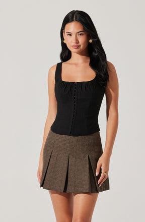 A woman wearing a sleeveless black top with a square neckline and button details down the front, paired with a high-waisted plaid skirt featuring pleats.