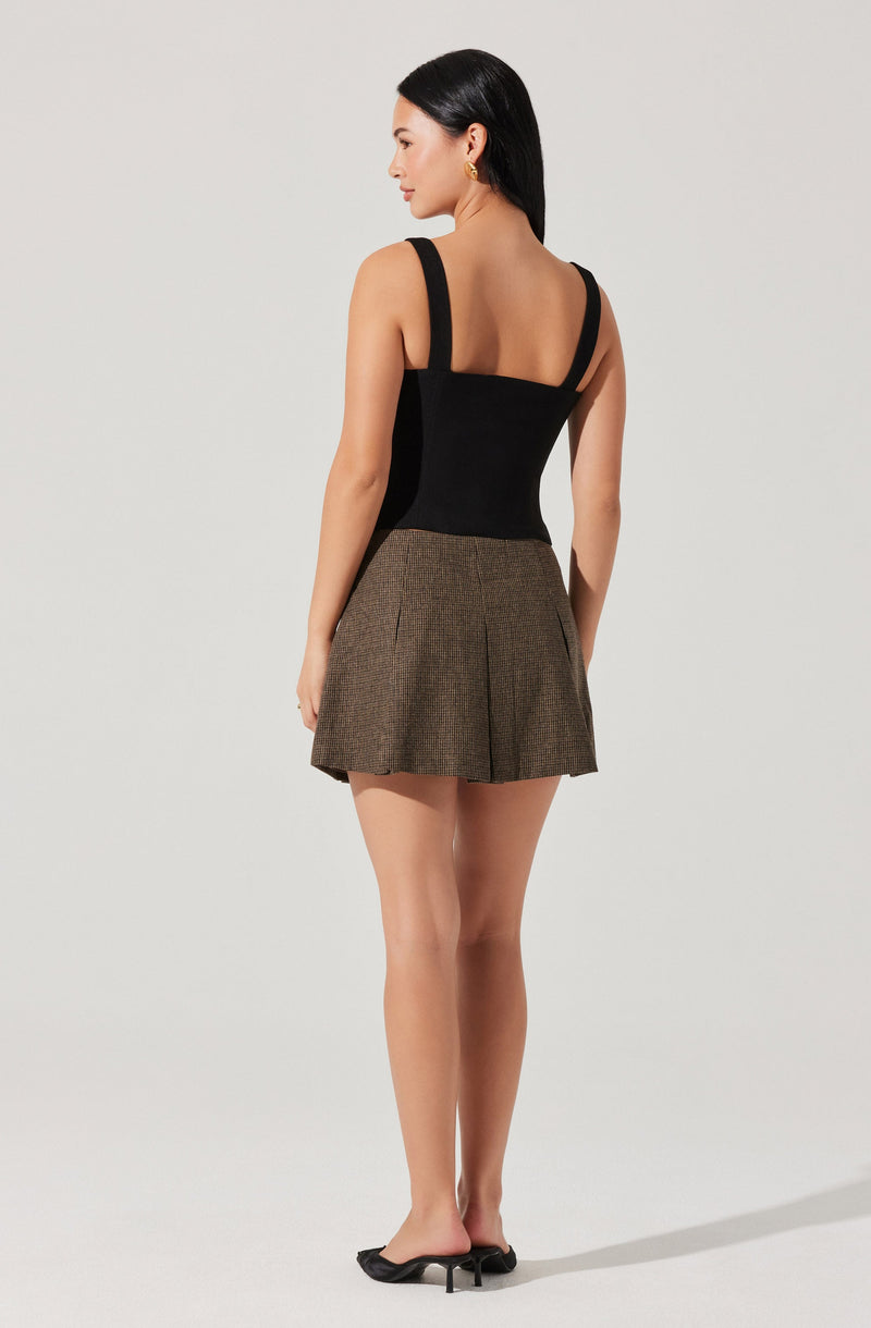 A woman is wearing a black sleeveless top with wide straps and a fitted bodice, paired with high-waisted, brown plaid shorts featuring pleats. She is also wearing black high-heeled shoes.