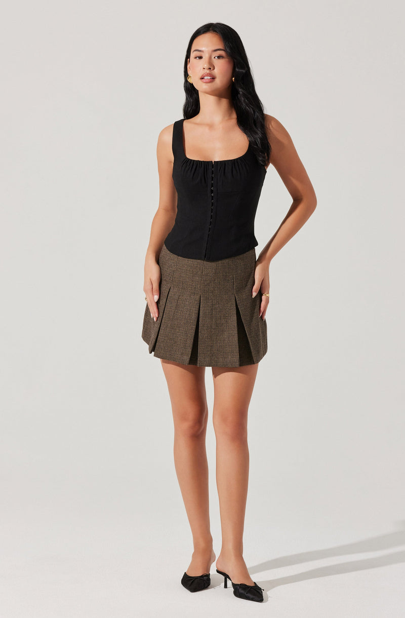 A woman is wearing a sleeveless black top with a square neckline and gathered fabric at the bust, paired with a high-waisted, brown plaid pleated skirt that has large pleats and a subtle pattern. She completes her outfit with black pointed-toe heels featuring a bow detail.
