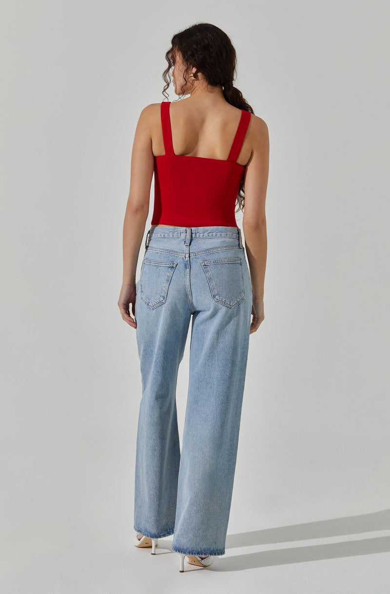 A woman is wearing a sleeveless red top with wide straps and a square neckline, paired with high-waisted, light-wash, wide-leg jeans. The jeans feature large back pockets and are paired with white high-heeled shoes. The woman has long, curly dark hair and is standing against a plain, light-colored background.