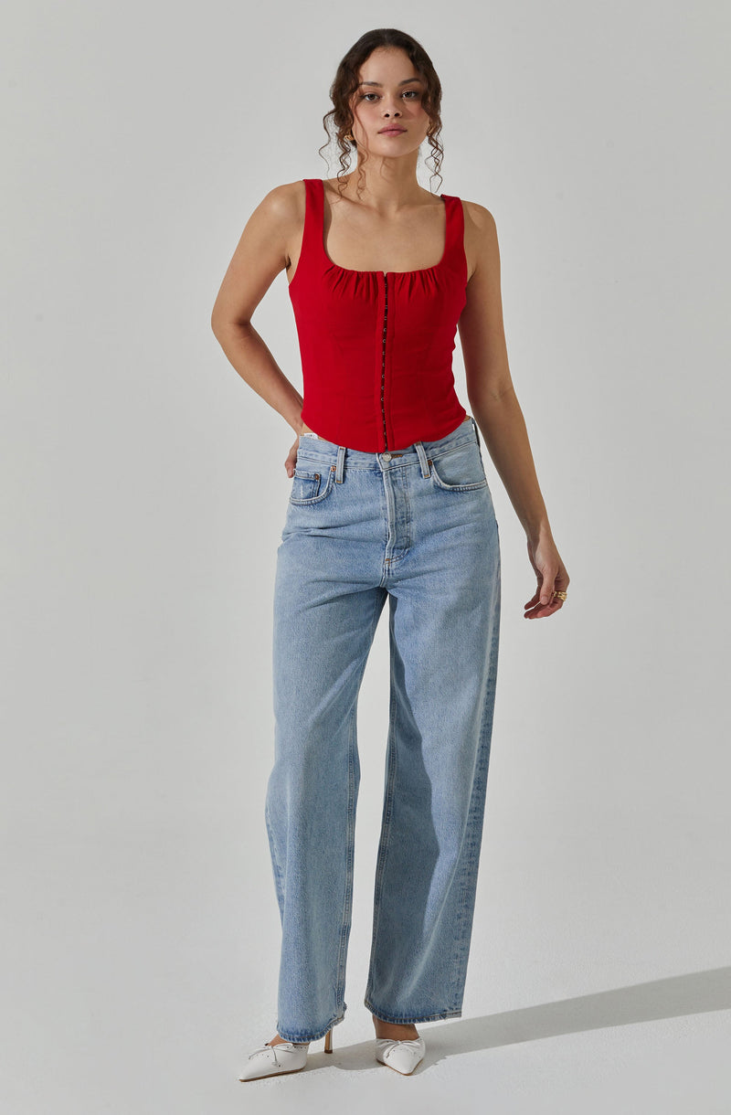 A woman is wearing a sleeveless red top with a gathered neckline and front zipper detail. She has paired it with light blue high-waisted, straight-leg jeans and white pointed-toe heels.