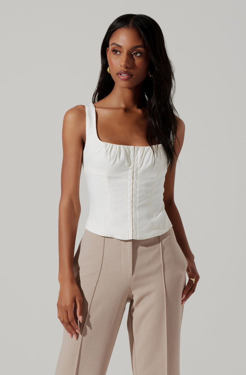 A woman is wearing a sleeveless, cream-colored top with wide straps and a square neckline, featuring a gathered detail along the neckline and a vertical seam down the center. She pairs it with high-waisted, light beige trousers that have a tailored fit with visible vertical seam lines on the front.
