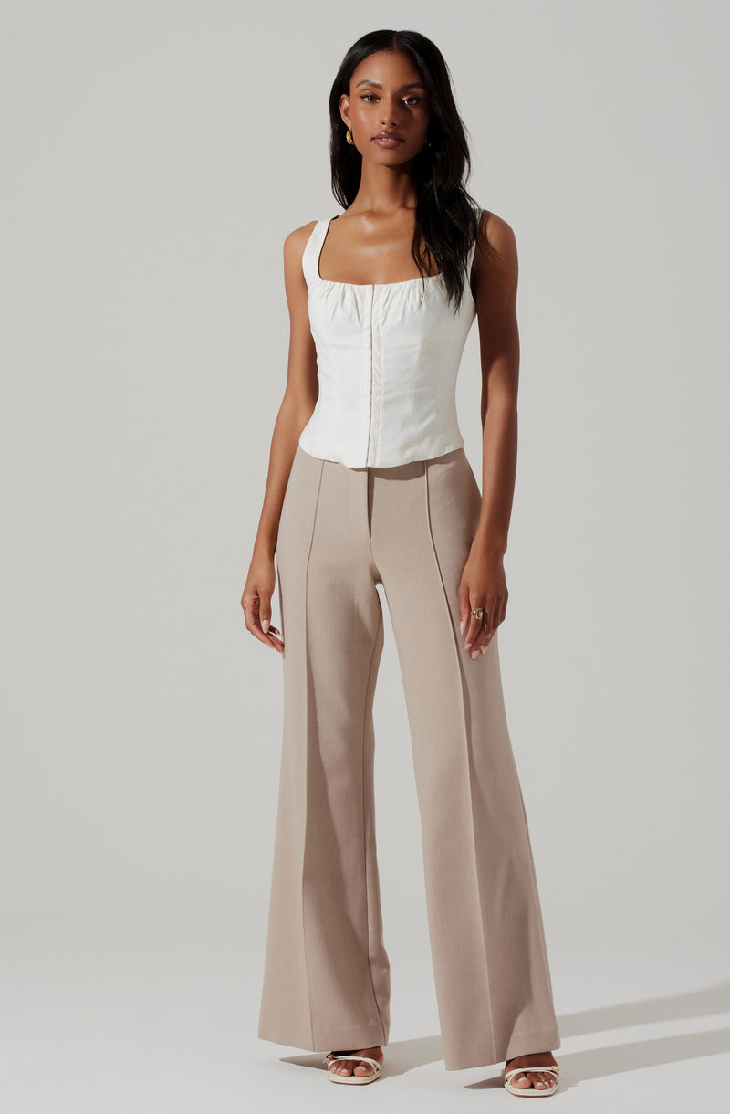 A woman is wearing a sleeveless white top with wide shoulder straps and a fitted bodice, paired with high-waisted, beige wide-leg trousers featuring vertical seam details.