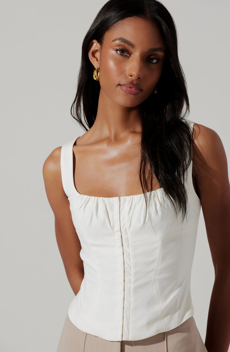 A woman is wearing a sleeveless, cream-colored top with a square neckline and gathered fabric detail at the bust. The top is fitted and has vertical seams running down the front. She has accessorized with gold hoop earrings and has long, dark hair styled in loose waves.