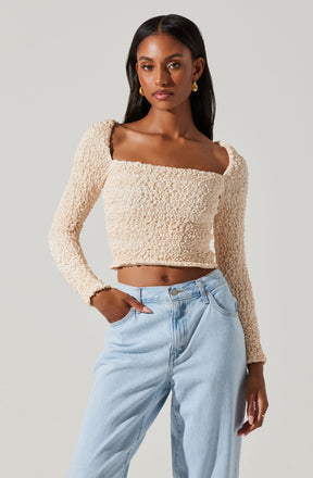 A woman is wearing a beige, textured, long-sleeve, crop top with a square neckline paired with light-wash high-waisted jeans. She accessorized with gold hoop earrings.