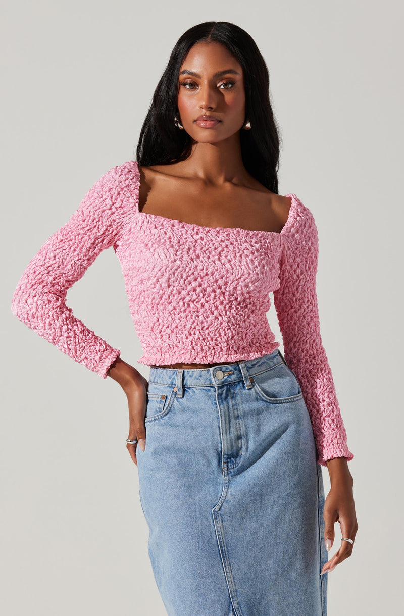 A woman is wearing a long-sleeved, textured pink top with a square neckline paired with a high-waisted, light-wash denim skirt.