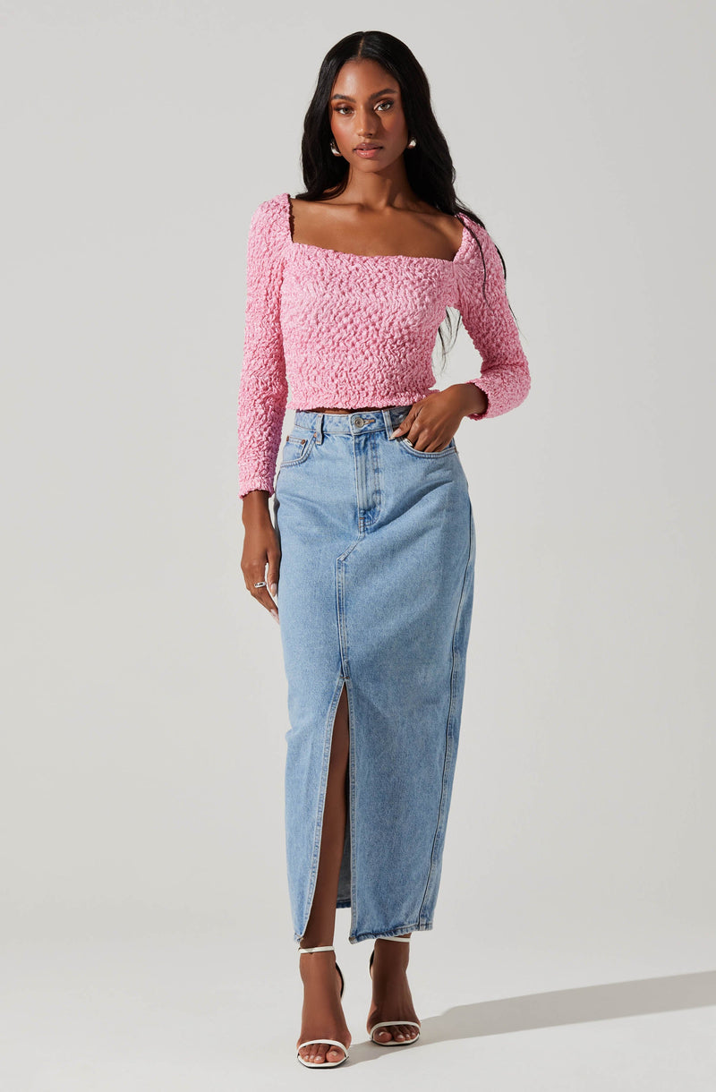 A woman is wearing a pink textured long-sleeve crop top with a wide square neckline, paired with a high-waisted, light blue denim maxi skirt that has a front slit. She is also wearing white high-heeled sandals.