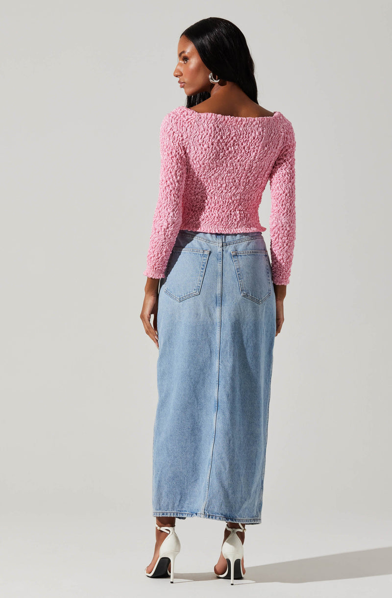 A woman is wearing a textured, long-sleeved pink sweater with a rounded neckline and a light-wash, high-waisted, ankle-length denim skirt with large back pockets. She is also wearing white high-heeled sandals with thin ankle straps.