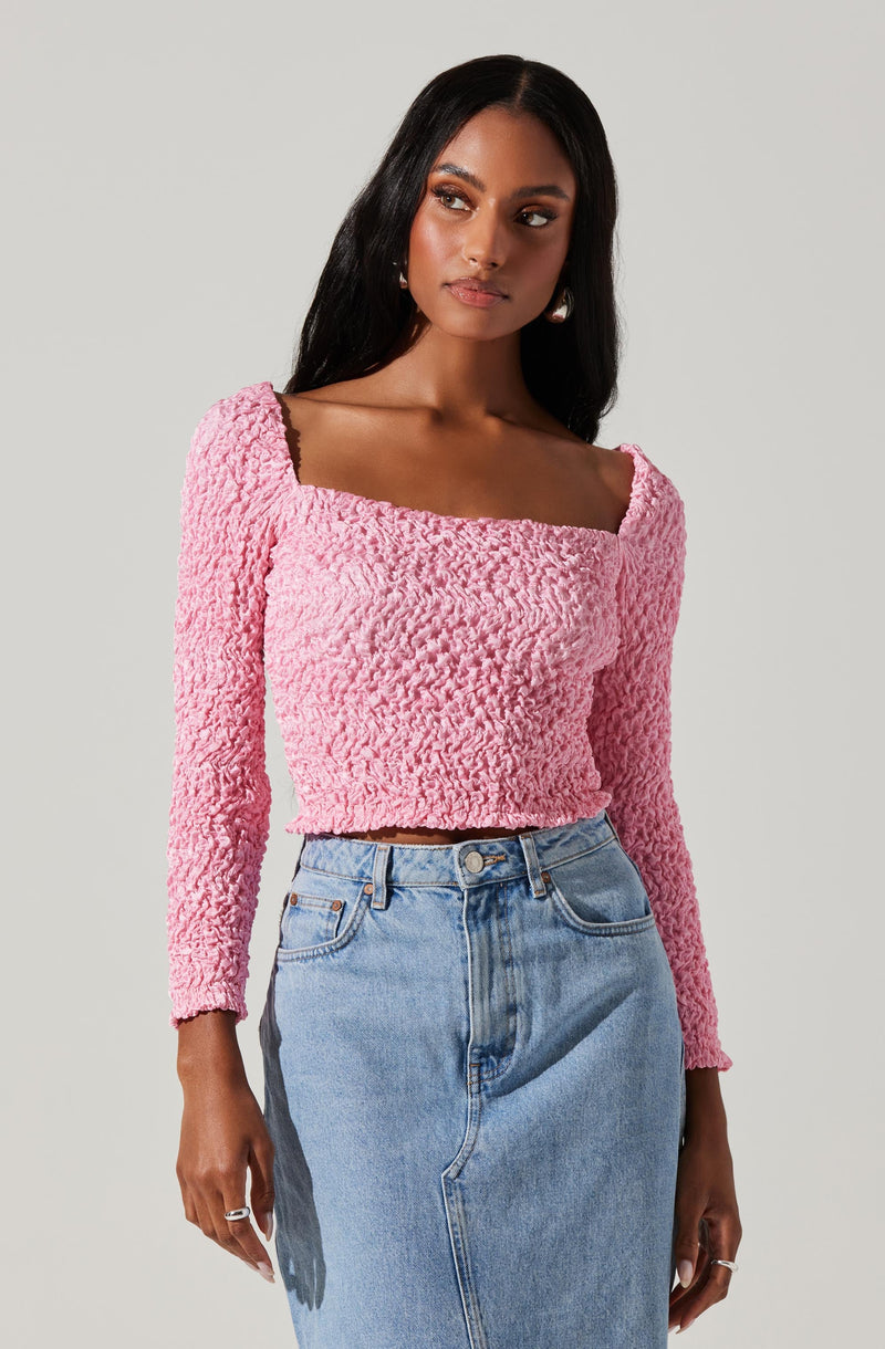 A woman is wearing a pink textured long-sleeve crop top with a square neckline and a light blue high-waisted denim skirt.