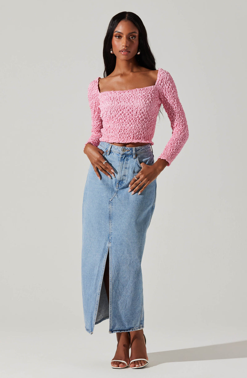 A woman wearing a pink textured long-sleeve top with a square neckline and puffed shoulders, paired with a high-waisted light blue denim maxi skirt with a front slit. She is also wearing open-toed heels.