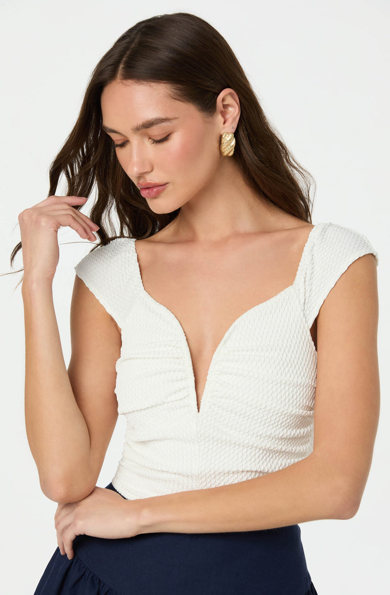 A woman is wearing a white textured top with a deep V neckline and cap sleeves, paired with dark high-waisted bottoms. She is accessorized with large, round, gold earrings.