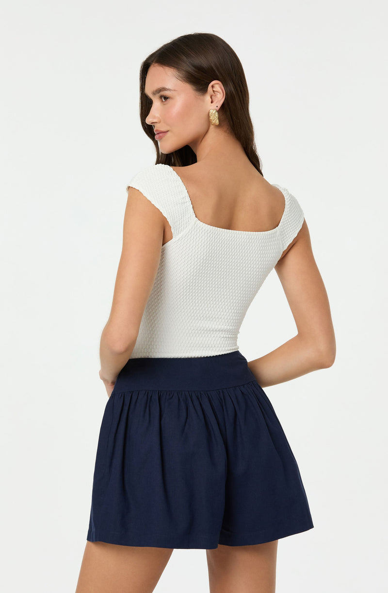 A woman is wearing a white, textured sleeveless top with a square neckline and shoulder straps, paired with high-waisted navy blue shorts that have a gathered, loose fit. She accessorizes with large gold earrings.