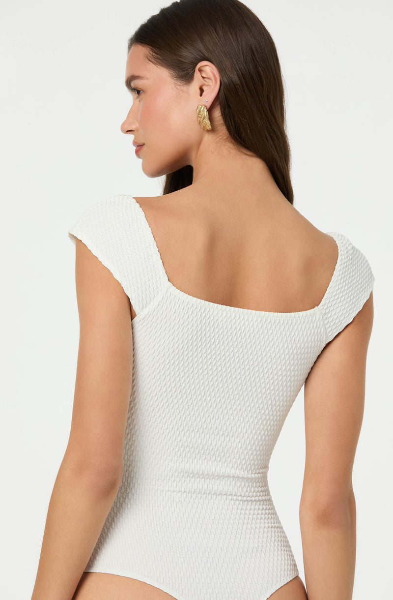A woman with long brown hair is wearing a white textured sleeveless top with a scoop neckline. She is accessorized with large, gold, textured hoop earrings. The background is plain and light-colored.