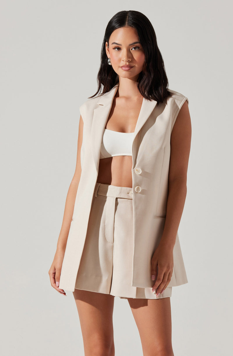 The woman is wearing a sleeveless cream-colored blazer with a notched collar and two buttons, paired with matching high-waisted shorts that have pleats and a belt loop. Underneath, she has on a white cropped top.