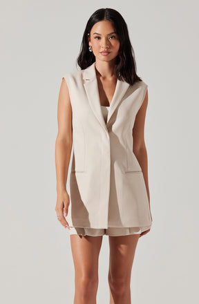 A woman is wearing a sleeveless, beige, tailored vest with lapels and two front pockets. She has paired it with matching beige shorts.