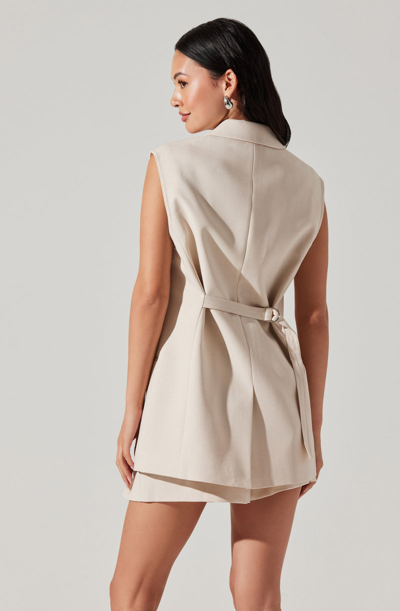 A woman is wearing a sleeveless, beige, tailored vest with a belt tied at the waist. The vest has a structured collar and side slit pockets. She is also wearing a matching beige skirt underneath.