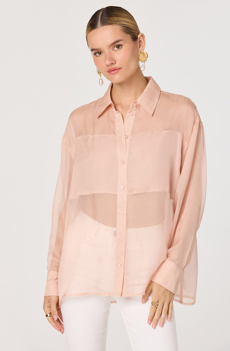 A woman is wearing a loose-fitting, long-sleeved, button-down shirt in a soft, blush pink color. The shirt is semi-transparent, revealing a darker blush or peach-colored camisole underneath. She has paired it with white pants. She accessorizes with large, gold, textured hoop earrings and multiple gold rings.