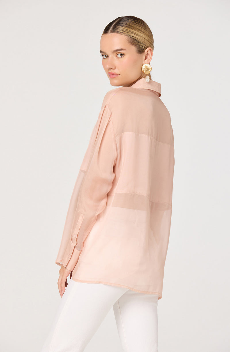A woman is wearing a semi-sheer, light pink, long-sleeved blouse with a collar. The blouse is loose-fitting and slightly oversized. She has paired it with white pants. Her hair is pulled back, and she is wearing large, circular, gold earrings with a decorative design.