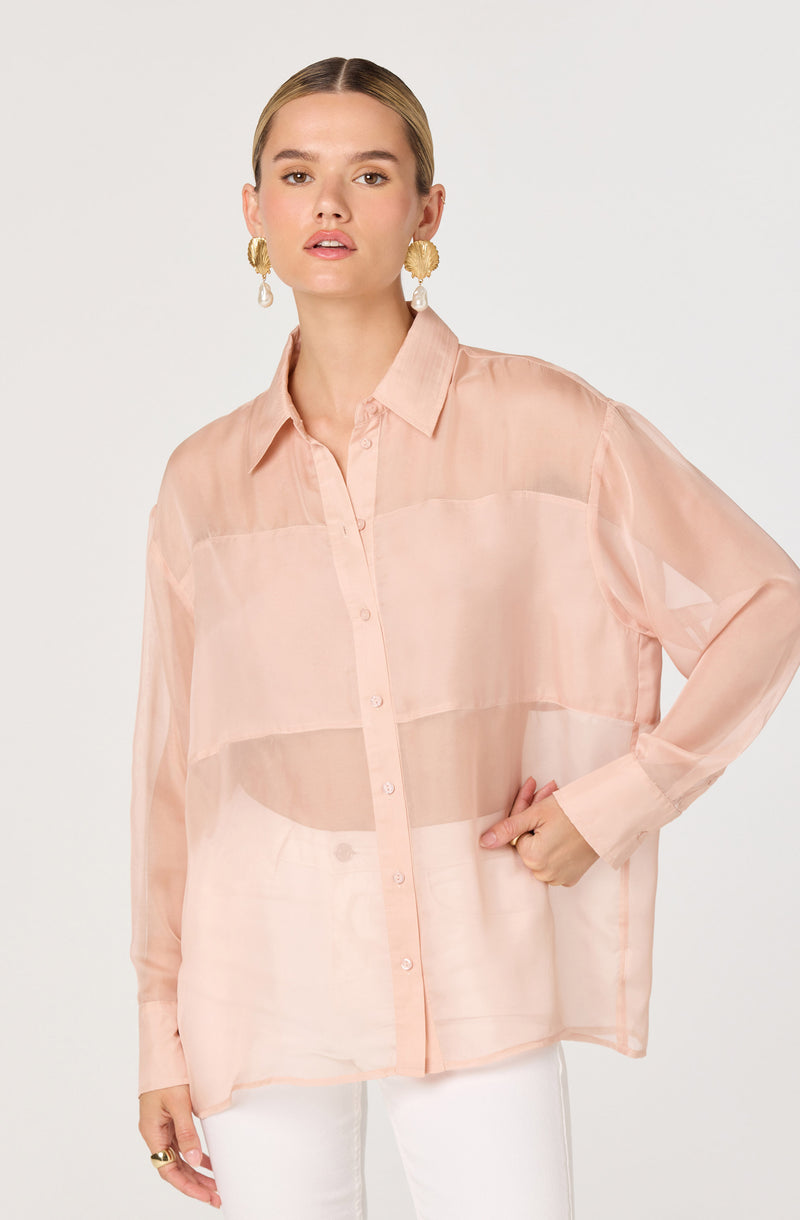 The model is wearing a loose, long-sleeved, button-up shirt made of a sheer, lightweight fabric in a soft, pale peach color. The shirt features a classic collar and vertical row of buttons down the front. She is also wearing large, gold, textured earrings with white, pearl-like pendants.