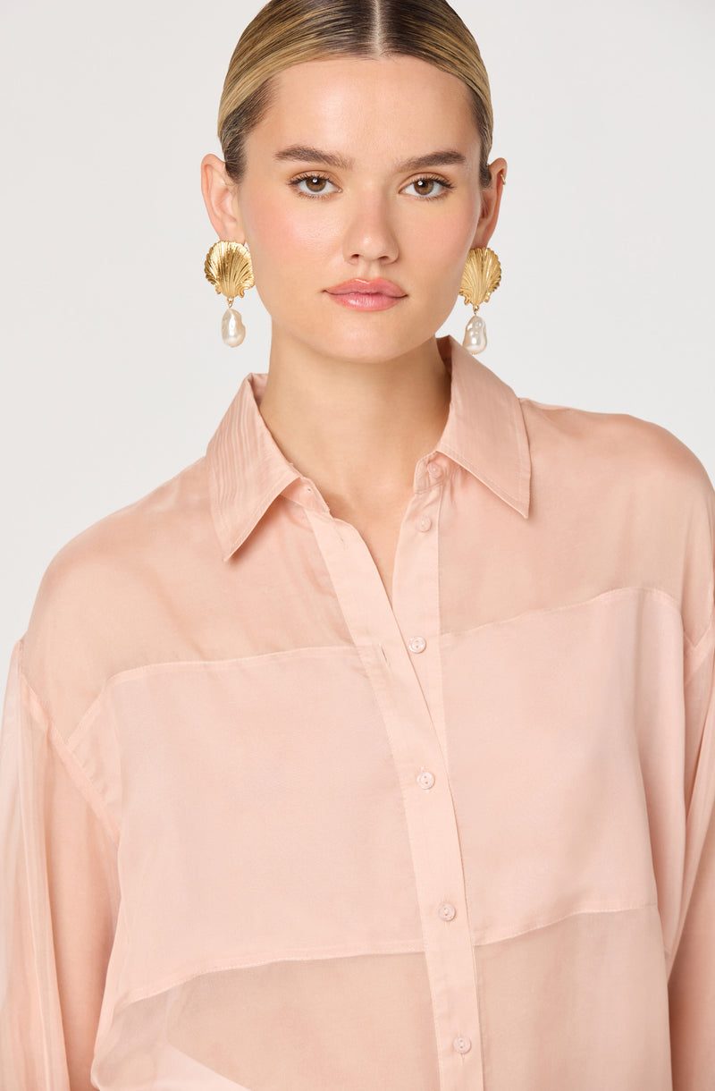 A woman is wearing a light pink, semi-sheer button-up blouse with a classic collar. The blouse is loose-fitting and appears to be made of a soft, flowing fabric. She has on large, gold-toned earrings that feature a shell design with dangling pearl-like accents.