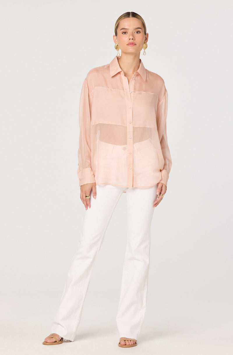 A woman is wearing a light pink, sheer, button-up blouse with long sleeves and a classic collar. She has paired it with white, straight-leg pants. She is accessorized with large gold hoop earrings and rings on her fingers, and she is wearing open-toed sandals.