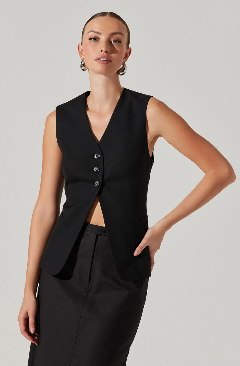 A woman is wearing a sleeveless black vest with a deep V-neck and four buttons down the front. The vest is fitted and tailored, with a slightly flared hem. She pairs it with a matching black skirt that has a high waist and a sleek, straight silhouette. She also has large, statement earrings.