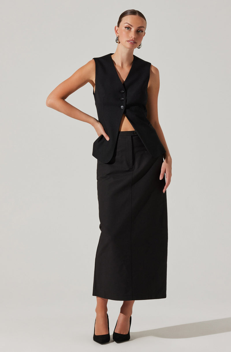 A woman is wearing a sleeveless black vest with buttons down the front, paired with a high-waisted, long black skirt that reaches her ankles. She completes the outfit with black high-heeled shoes.