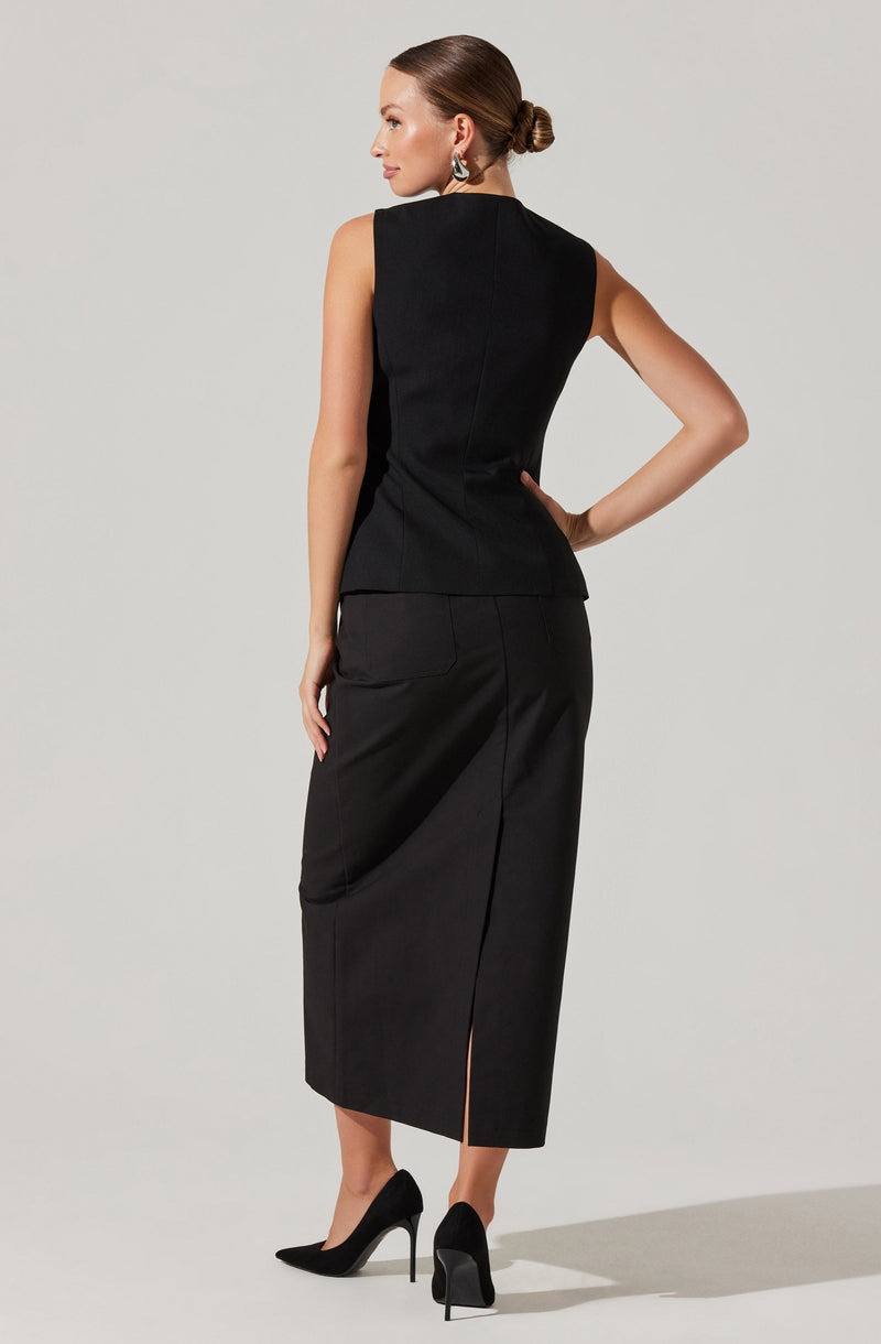 A woman is wearing a sleeveless, fitted black top with a high neckline, paired with a long, black, high-waisted skirt that features a front slit. She is also wearing black high-heeled shoes and has her hair styled in a sleek, low bun. She accessorizes with earrings.