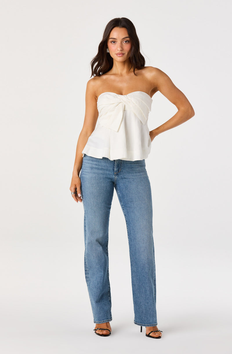 Woman wearing a white strapless top with a large bow detail and ruffled hem, paired with blue jeans.