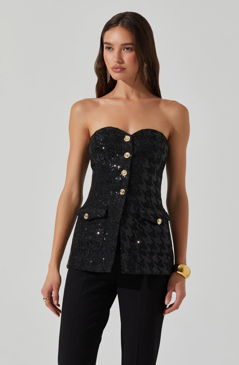 A woman is wearing a strapless black top with sequins and decorative buttons down the front. The top features a textured, patterned fabric on one side and a smooth, sequined surface on the other. She pairs it with black pants and accessorizes with large earrings, a gold watch, and a ring.