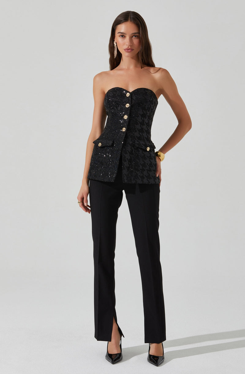 The woman is wearing a strapless black top that features a textured pattern and decorative gold buttons down the front, with a small pocket on the right side. She pairs the top with tailored black pants that have a slit at the hem. She is also wearing black pointed-toe heels and a gold bracelet on her right wrist.
