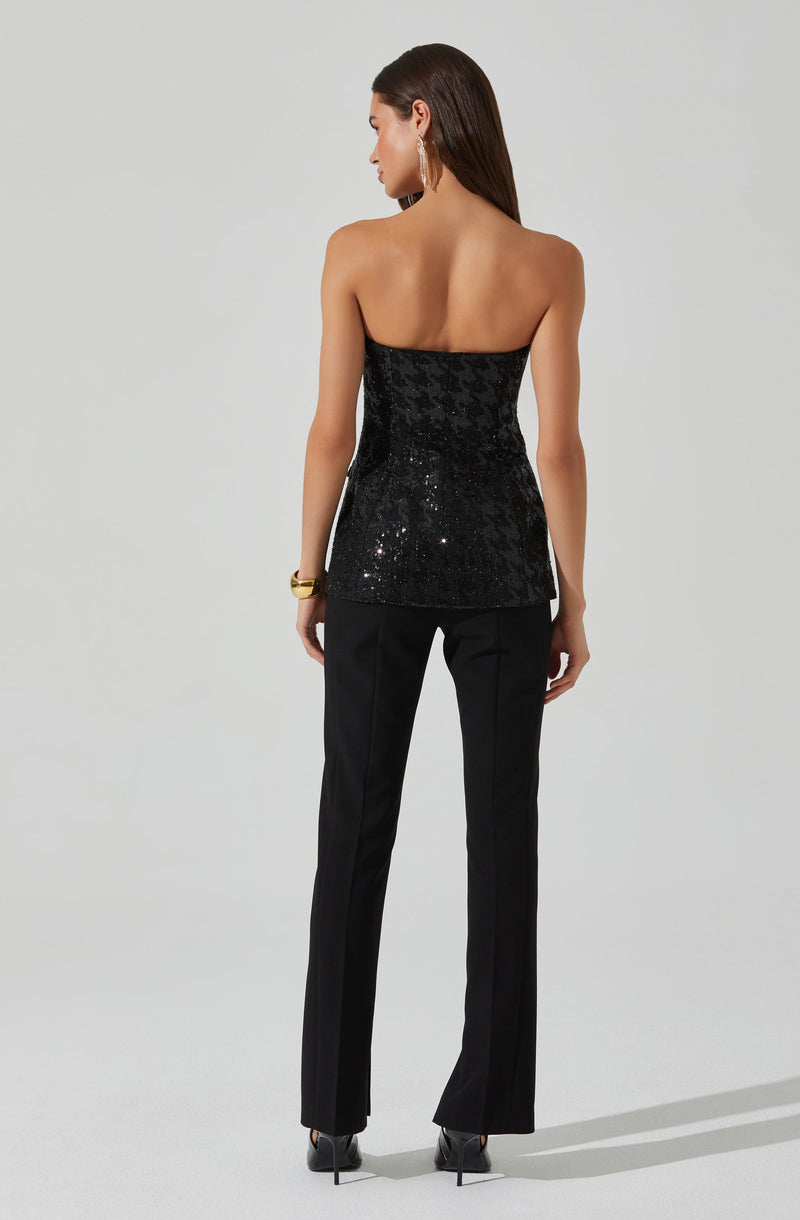 A woman is wearing a strapless black top with sequins that create a shiny, textured appearance. She pairs it with tailored black pants that have a slim fit through the hips and thighs, flaring slightly at the hem. She accessorizes with large hoop earrings, a gold bracelet on her left wrist, and black high-heeled shoes. Her long dark hair is worn down, and she stands against a plain, light-colored background.