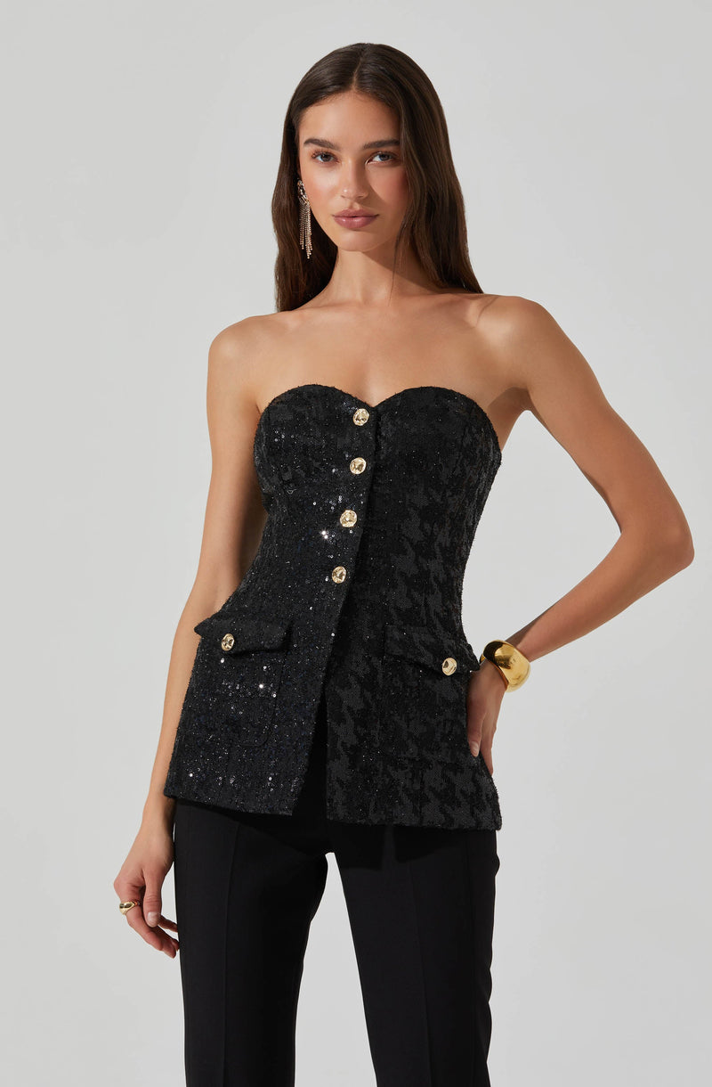 A woman is wearing a strapless black top with a textured, velvet-like pattern and a row of large, decorative gold buttons down the front. The top features a pocket with a flap on the right side, also adorned with a gold button. She accessorizes with long, dangling earrings and a wide, gold bracelet on her left wrist.