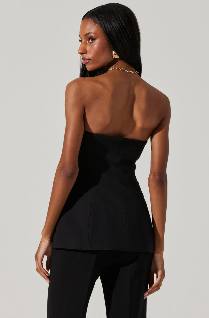 A woman is wearing a strapless black fitted top and matching black pants. The top is tailored and form-fitting, with a flared hemline at the waist. She accessorizes with gold earrings and a gold chain necklace.