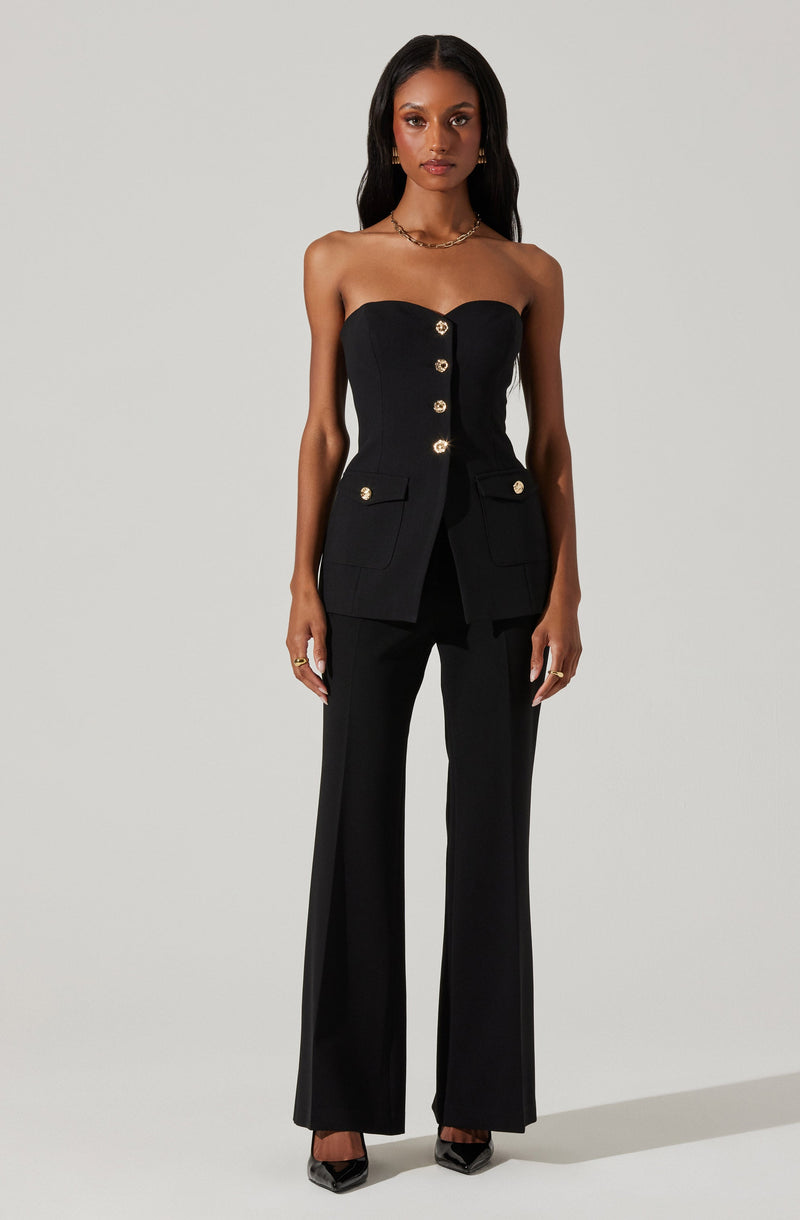 The outfit consists of a strapless black top with a structured, sweetheart neckline, featuring gold buttons down the center and on the pockets. The top is fitted at the waist and is paired with matching high-waisted black wide-leg pants. The ensemble is complemented by black pointed-toe shoes and gold jewelry.