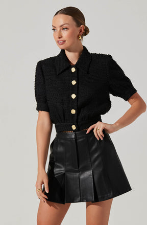 A woman is wearing a black, textured, short-sleeved button-up top with a collar and large, gold buttons down the front. She pairs it with a high-waisted, black leather mini skirt featuring vertical pleats.