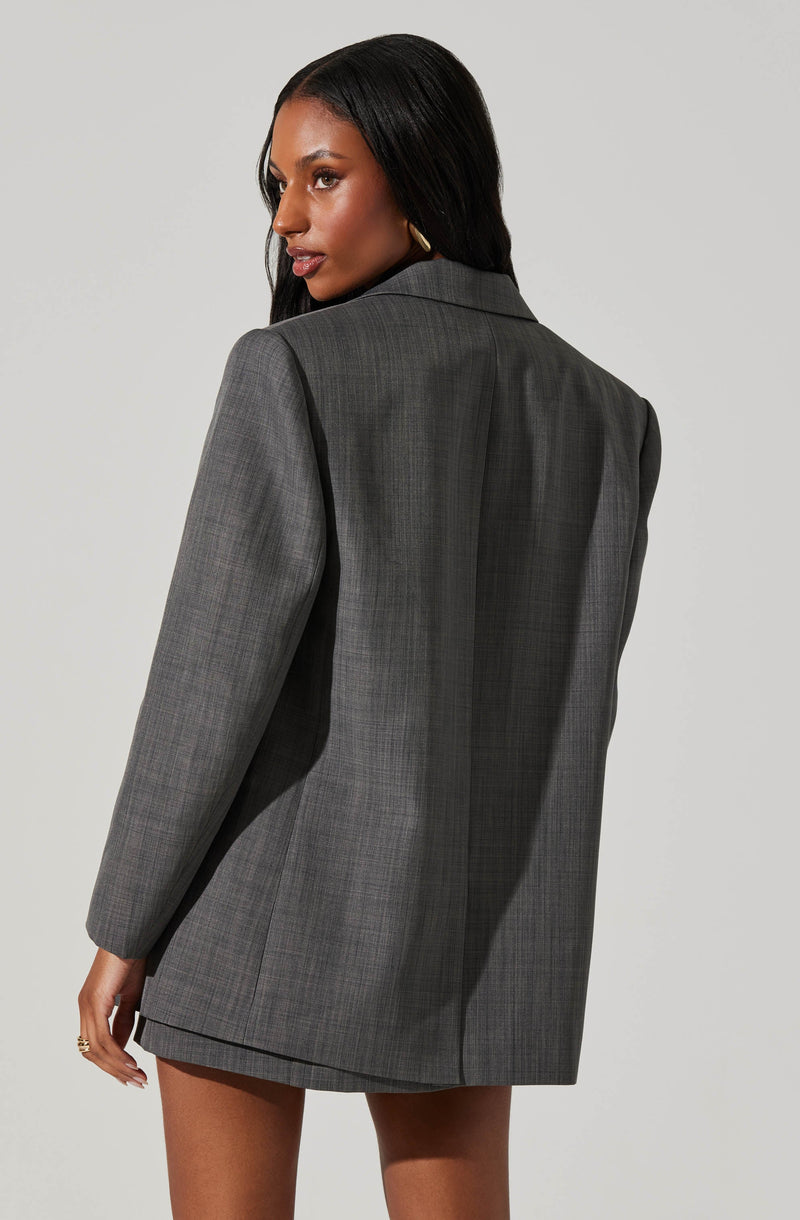 A woman is wearing an oversized, gray plaid blazer with a classic lapel collar. The blazer features a subtle checkered pattern, structured shoulders, and long sleeves, and it extends to mid-thigh length.