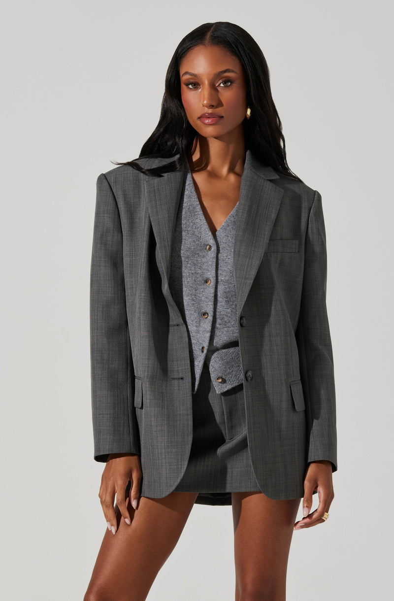 A woman is dressed in a gray checked blazer with structured shoulders, paired with a matching gray cardigan and skirt underneath. The blazer has a classic notch lapel, and the cardigan features small buttons down the front. The skirt is matching in color and pattern, creating a coordinated, professional look.
