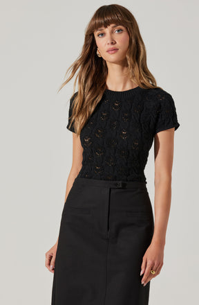 A woman is wearing a black short-sleeved knit top with a delicate, open weave pattern. She has paired it with a high-waisted black skirt that features a front zipper closure and a small belt loop.