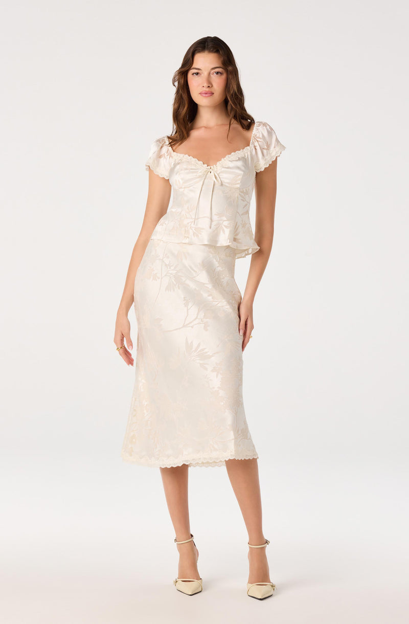 Woman wearing a cream satin top with ruffled sleeves and a matching midi skirt with a floral pattern and scalloped hem.