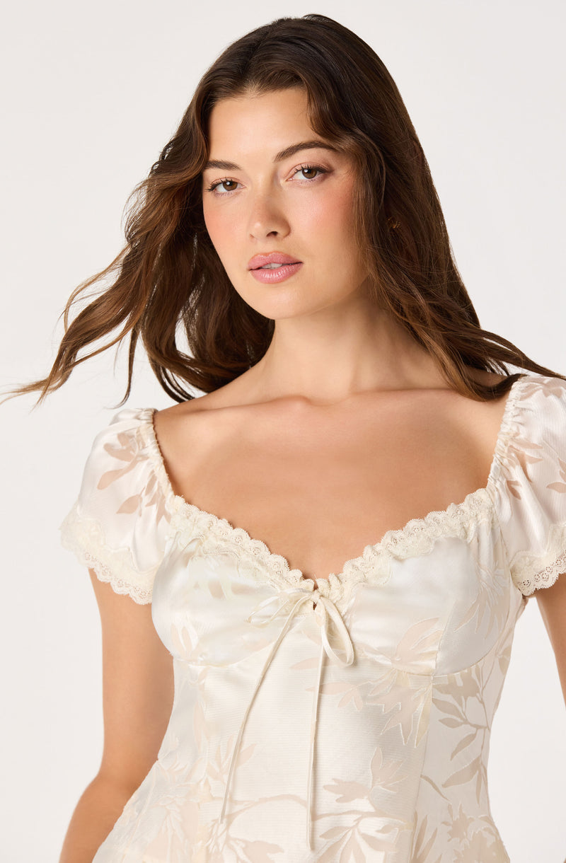Woman wearing a cream satin dress with lace trim and a front bow detail.