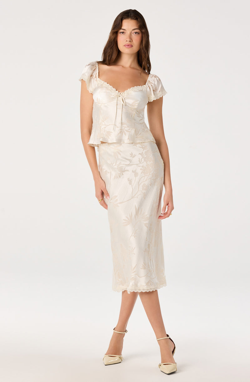 Woman in cream satin dress with floral pattern, puffed short sleeves, and bow detail.