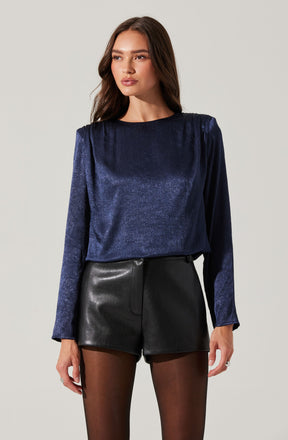 A woman is wearing a long-sleeved, shiny, cobalt blue velvet blouse with a high round neckline. She pairs it with high-waisted black leather shorts and sheer black tights.