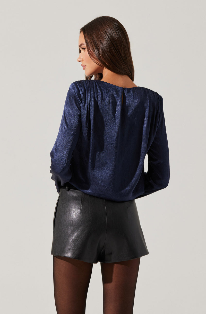 A woman is wearing a dark blue, long-sleeved, shiny blouse with gathered fabric details on the shoulders and upper back. She is also wearing high-waisted black leather shorts and sheer black tights.